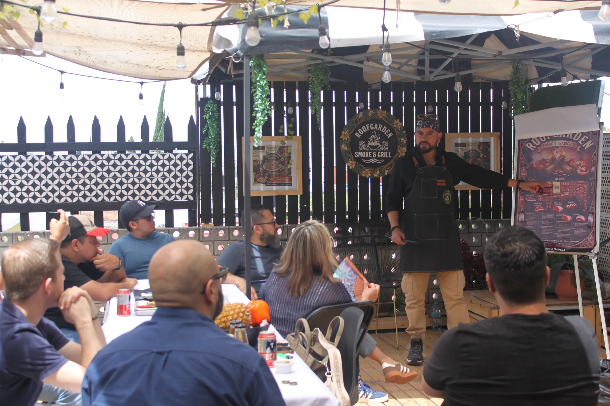Chef instructor de RoofGarden explicando cortes de carne frente al grupo, junto al letrero RoofGarden Smoke & Grill